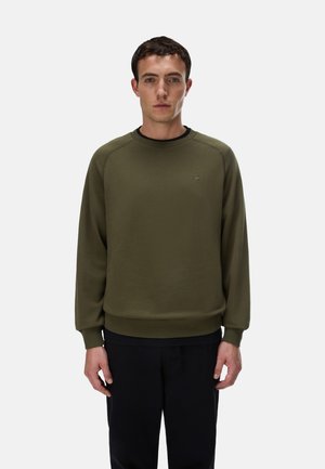Jeune homme aux cheveux courts et foncés portant un sweat-shirt vert olive à col rond et un pantalon noir, debout devant un fond clair uni.