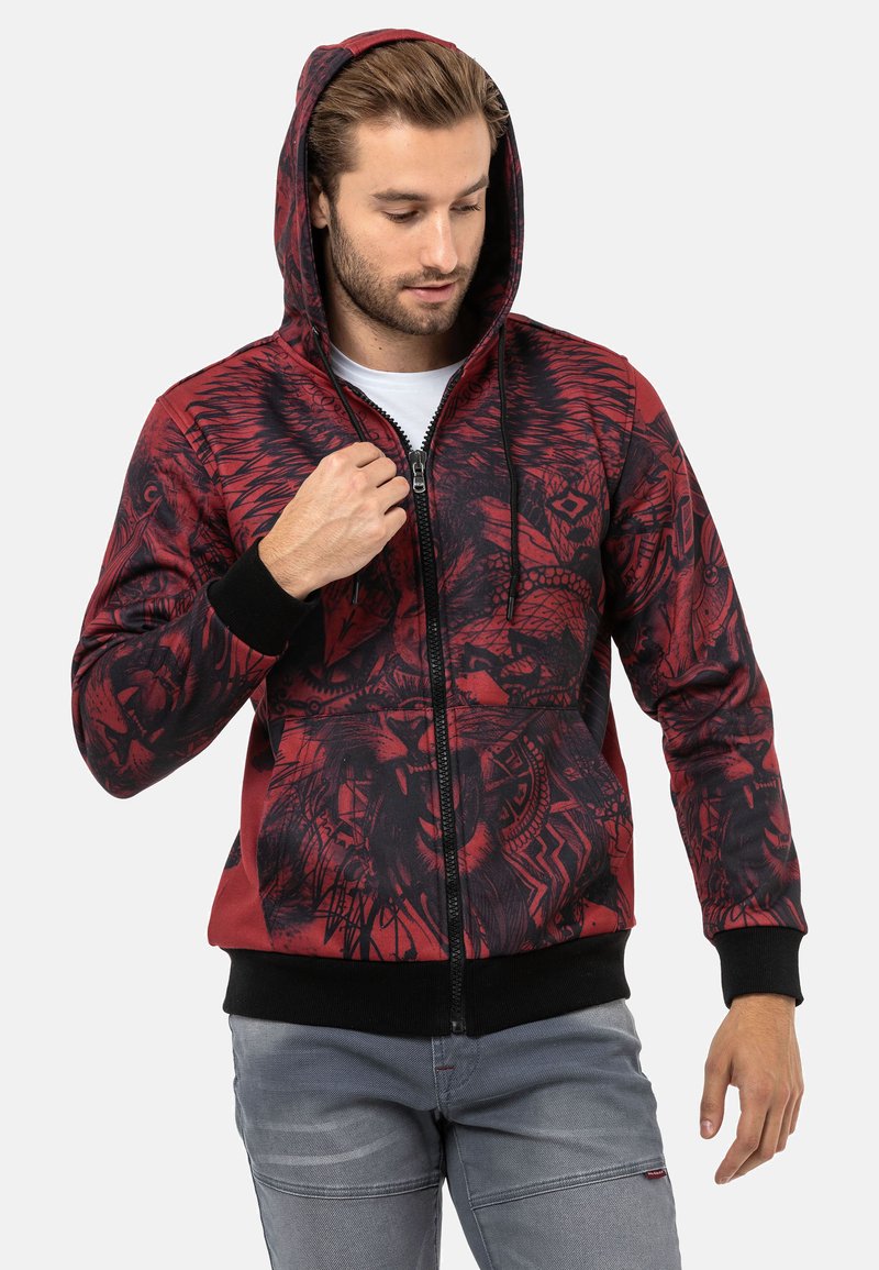 Rote und schwarze Zip-Hoodie mit einem grafischen Design, das Löwen und Tribal-Muster zeigt. Verfügt über eine Kapuze mit Kordelzug und ribbelte Bündchen.