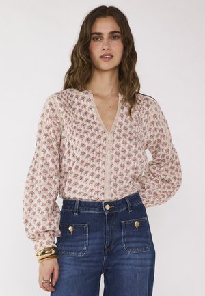 Femme aux cheveux bruns ondulés portant un chemisier à manches longues rose clair à motifs floraux et un jean bleu taille haute avec poches avant, bracelets dorés au poignet gauche.