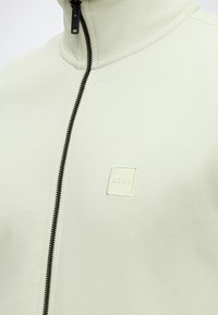 Κοντινό πλάνο ανοιχτό μπεζ τζάκετ με φερμουάρ σε μαύρο χρώμα και ανάγλυφο λογότυπο BOSS στο αριστερό στήθος.