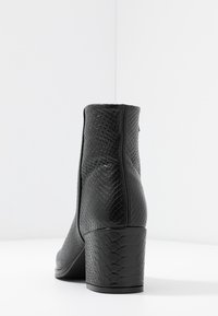 Černá kotníková bota s blokovým podpatkem, zdobená texturou hadí kůže, zobrazená zezadu na hladkém bílém povrchu a pozadí.