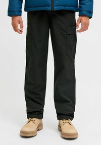 Personne portant un pantalon cargo noir avec des poches latérales, des bottes beiges à lacets et une veste en doudoune bleue partiellement visible au niveau de la taille.