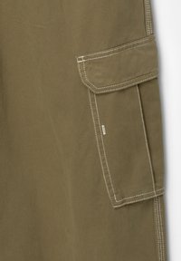 Dettaglio di pantaloni in tessuto kaki che mostra la tasca laterale cargo con patta e cuciture bianche a contrasto su materiale di cotone resistente.