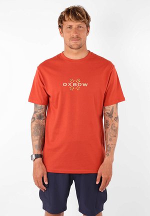 T-shirt en coton rouge avec un logo central "Oxbow" orné de feuilles. Manches courtes et encolure ras du cou classique. Texture lisse.
