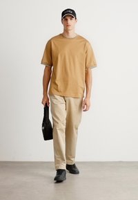 Mann som går iført en lys t-skjorte, beige bukser, svarte joggesko, svart caps og bærer en liten svart veske i venstre hånd.
