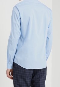 Light blue, long-sleeve shirt with a smooth texture, button cuffs, and a curved hem. Worn with dark plaid pants, showing a relaxed fit.