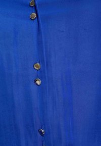 Camicia di stoffa blu con una texture liscia e un taglio dritto, caratterizzata da cinque bottoni metallici rotondi lungo la parte anteriore.