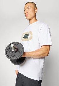 Czarny kettlebell z metalowym uchwytem i plastikowymi chwytami. Model ma na sobie białą bawełnianą koszulkę z zielono-beżowym logo.