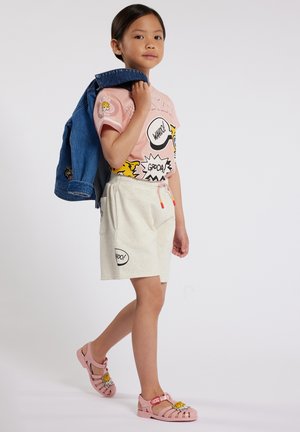 Een kind in een roze grafisch T-shirt met print in stripstijl, witte gemarmerde shorts en roze sandalen met cartoonaccenten, terwijl het een spijkerjack vasthoudt.