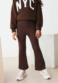 Ein Kind trägt einen braunen Sweatshirt mit "NYC" Grafik, kombiniert mit braunen, ausgestellten Leggings und rosa Turnschuhen, in einer minimalistischen, hellen Umgebung.