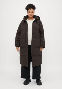 Dark brown padded coat with a hood, featuring a front zipper, press-stud closures, and side pockets. Worn over a light sweater and pants.