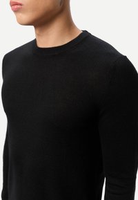 Pull en tricot noir avec col rond, coupe ajustée, texture douce et tissu lisse, doté de manches longues et d'un design épuré et minimaliste.