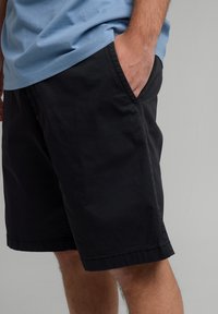 Black cotton shorts with side pockets, straight cut, and a stitched hem. Paired with a light blue shirt, showing a relaxed fit.