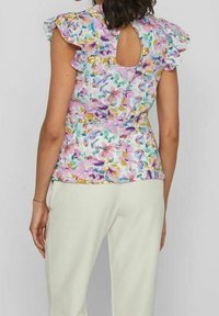 Femme portant un chemisier sans manches à volants et à motifs floraux, avec un détail en forme de trou à l'arrière, assorti à un pantalon blanc taille haute.