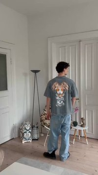 T-shirt gráfica oversized cinza com impressão nas costas, combinada com jeans de denim azul claro. Calçado preto simples e acessórios mínimos.