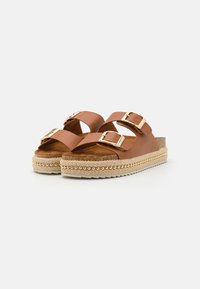 Sandalias de cuero marrón tipo chancla con dos correas ajustables y hebillas doradas, una gruesa plataforma de esparto trenzada y una suela texturizada.