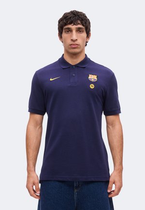 Homme portant un polo bleu marine avec un logo Nike jaune, le blason du FC Barcelone et le logo Spotify, debout devant un fond clair uni.