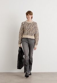 Beige Pullover mit Leopardenmuster, gerippten Ärmelbündchen, kombiniert mit grauen Schlagjeans und schwarzen Stiefeletten, hält eine schwarze Tragetasche.