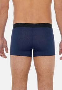 Navy blue trunks with a smooth fabric texture, featuring a black elastic waistband displaying the logo. Cut to mid-thigh length.