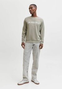 Grå sweatshirt med "JACK & JONES" logo, kombineret med lyse slidte jeans og sølvfarvede sneakers, stående mod en ensfarvet baggrund.