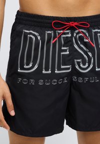 Mustad ujumispüksid elastse vöökohaga ja punase paelaga. Silmatorkav hõbedane tekst: "DIESEL FOR SUCCESS." Siledapind ja püksid ulatuvad reie keskosani.