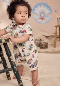 Shorty en tissu crème avec des motifs de tracteurs colorés. Conçu avec des poches et une fermeture à boutons, adapté aux tout-petits.