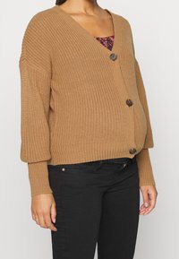 Donna che indossa un cardigan lavorato a maglia marrone con tre grandi bottoni sopra una maglietta a fantasia e pantaloni neri, mostrata dalla metà del petto ai fianchi.