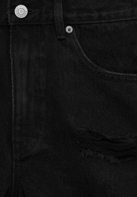 Gros plan d’un jean en denim noir montrant la poche avant, le bouton en métal, le rivet, la boucle de ceinture et une déchirure horizontale usée sous la poche.
