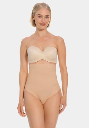 Woman with short blonde hair wearing strapless beige bra and high-waisted nude shapewear standing against plain light background.