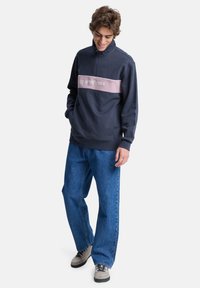 Navy Zip-Up-Fleece-Sweatshirt mit einem pinken horizontalen Streifen, der das Logo zeigt. Kombiniert mit lockeren blauen Jeans und beigen Turnschuhen.