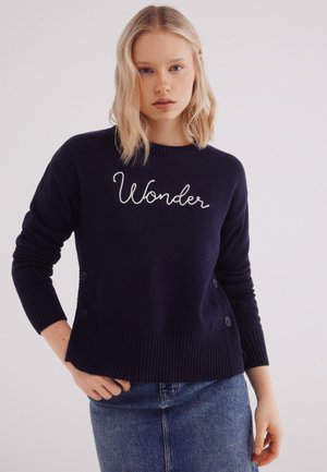 Marineblauwe gebreide trui met geribbelde zoom en boorden, voorzien van witte geborduurde tekst "Wonder" en knopenaccenten aan de zijkanten.