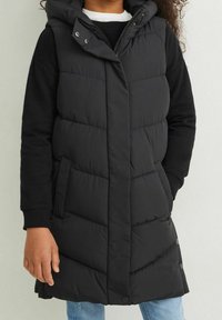 Czarna puchowa kamizelka bez rękawów z kapturem, z zapięciem na guziki, bocznymi kieszeniami i pikowanym wzorem. Noszona na czarnej koszuli z długim rękawem.