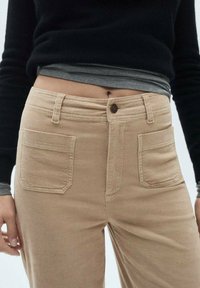 Beige corduroy-housut suoran lahkeen muotoilulla, joissa on kaksi etutaskua ja nappikiinnitys, näkyvät tekstuuriraidat läpi.