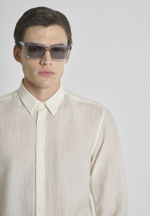 Jonge man met naar achteren gekamd donker haar, draagt donkere rechthoekige zonnebril en een crème kleurig, gestructureerd overhemd met knopen, tegen een effen achtergrond.