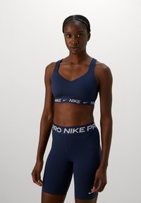 Persoană atletică îmbrăcată într-un sutien sport Nike albastru marin și pantaloni scurți de compresie cu talie înaltă asortați, pe un fundal gri simplu.
