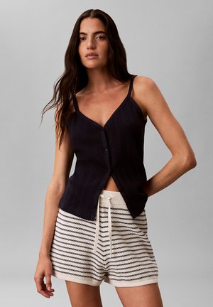 Femme portant un caraco noir boutonné et un short en maille blanc avec des rayures horizontales noires sur un fond gris uni.