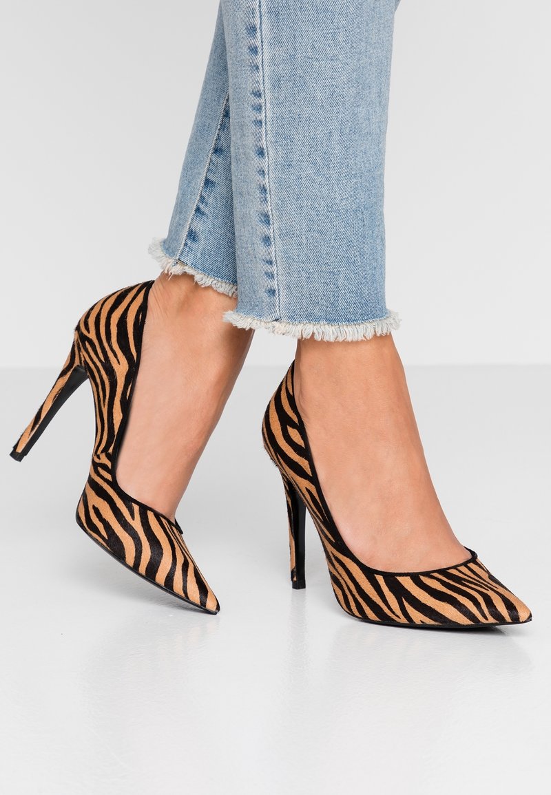 Pieds portant un jean bleu clair à ourlet effiloché et des escarpins à bout pointu avec des rayures tigre noires et marron.