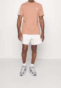 Ljus persikofärgad t-shirt med vit tre-stripes design på axlarna, i kombination med vita shorts och grå sportskor. Mjuk bomullsmaterial.