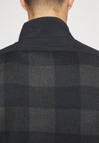 Vue de dos d'une personne portant une veste à carreaux noir et gris avec un col noir uni, montrant un gros plan sur la texture et les détails des coutures.