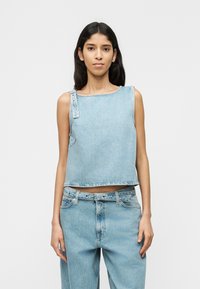 Jeune femme aux longs cheveux foncés porte un haut en denim bleu clair sans manches et un pantalon en denim taille haute assorti, sur un fond uni.