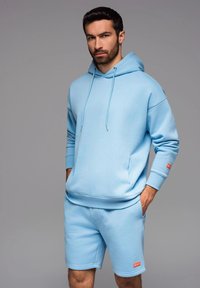Ljusblå hoodie med framficka och dragsnoddar, i kombination med matchande shorts. Mjuk tyg, avslappnad passform och liten röd logotypdetalj.
