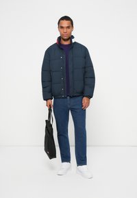 Chaqueta acolchada azul marino con cierre a presión, que presenta un cuello en contraste. Llevada sobre una camisa púrpura con jeans de mezclilla y zapatillas blancas, portando un bolso negro.