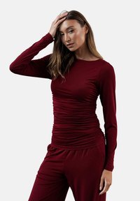 Burgundowa bluzka z długim rękawem i detalami marszczenia, wykonana z miękkiego materiału. Posiada okrągły dekolt i jest zestawiona z pasującymi spodniami.