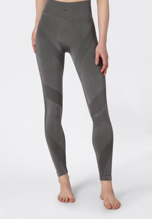 Donna che indossa leggings grigi a vita alta e senza cuciture con motivi a strisce diagonali scure, in piedi a piedi nudi su uno sfondo bianco.