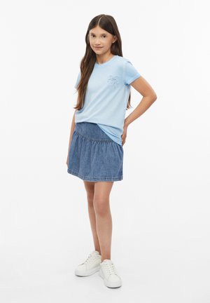 Mädchen mit langen braunen Haaren trägt ein hellblaues T-Shirt, einen Jeansrock und weiße Turnschuhe und steht mit einer Hand in der Hüfte vor weißem Hintergrund.