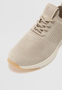 Beige Sport Schuh mit einem gestrickten Obermaterial, strukturiertem Muster, runder Zehenpartie, flachen Schnürsenkeln und einer leichten Gummisohle. Verfügt über ein Markenetikett auf der Zunge.