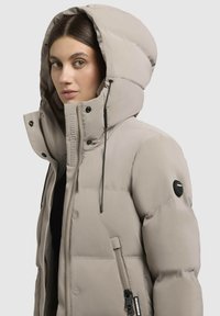 Pufjakas gaiši bēšā krāsā ar augstu apkakli un lielu kapuci. Iespējas redzamiem rāvējslēdzējiem un zīmola uzšuvēm uz piedurknes. Gluda tekstūra.