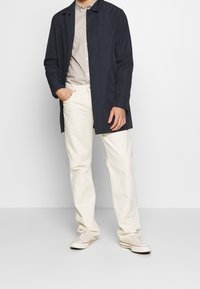 Marineblauwe lichtgewicht regenjas met een knoopsluiting, opstaande kraag, gedragen over een lichtgrijs shirt. Gepaard met crème witte broek en sneakers.