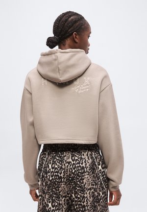 Kvinde med flettet hår iført en beige cropped hættetrøje med tekst og leopard print bukser, stående med ryggen til mod en ensfarvet baggrund.