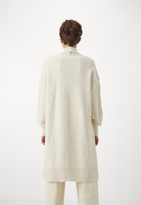 Osoba s krátkými kudrnatými vlasy, oblečená v oversize krémovém pleteném kardiganu a odpovídajících kalhotách, stojící zády k jednoduchému pozadí.